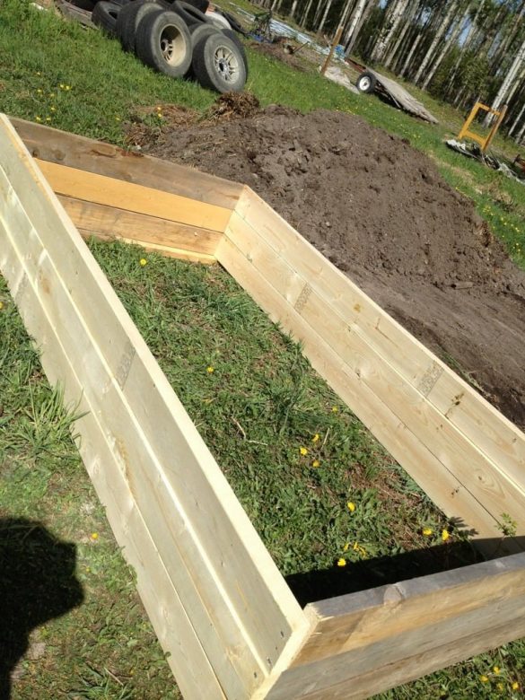 A wooden garden bed frame rests on grassy ground, beside a pile of soil. Tires and trees are silhouetted in the background, completing this rustic scene by Owl Creek Farm.