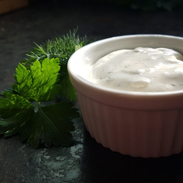 White bowl with creamy sauce by Owl Creek Farm sits next to fresh green herbs on a dark background.