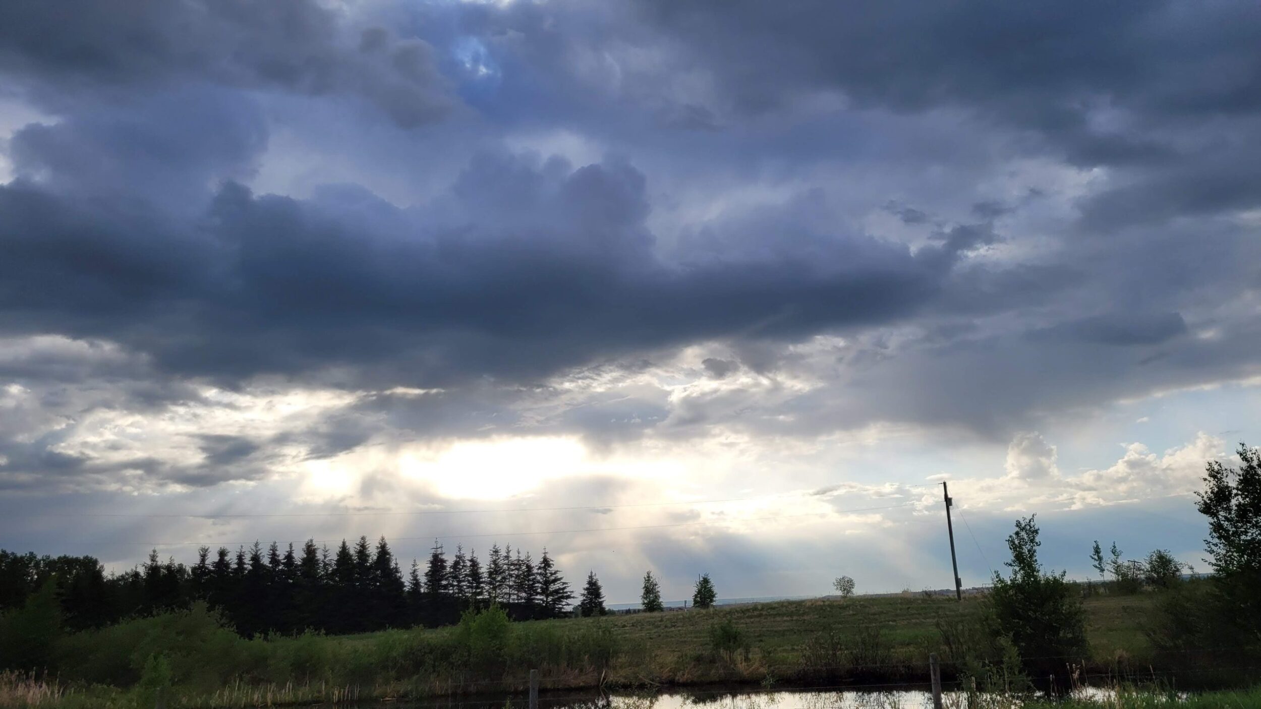 Photo of a cloudy sky with sun rays coming through the clouds, over a water filled dugout in Alberta. Owl Creek Farm